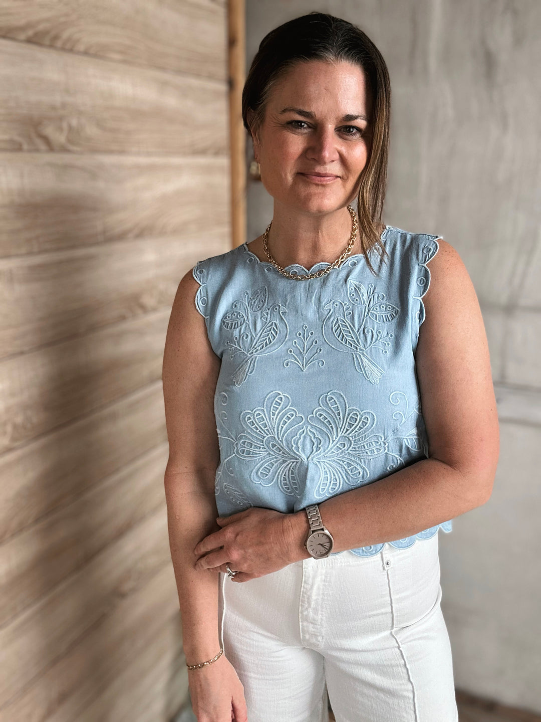 Woman wearing a light blue sleeveless top with embroidered details against a wooden wall.