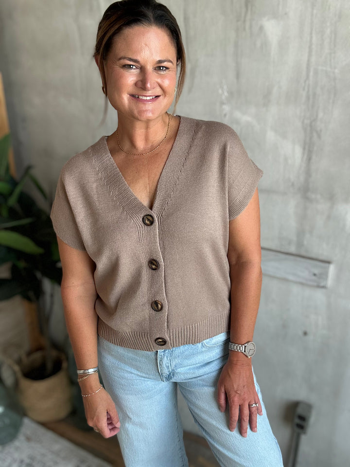 A woman wearing a taupe short-sleeve knit cardigan with dark buttons and light blue jeans, standing against a textured gray concrete wall.