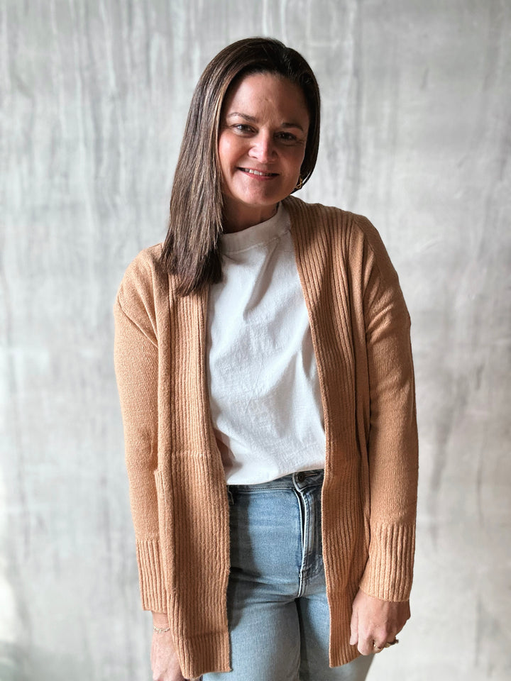 Woman wearing a beige cardigan over a white shirt and blue jeans against a plain background
