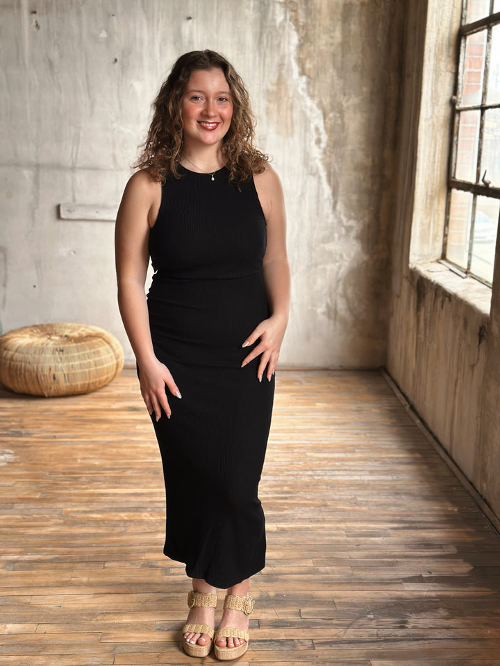 Woman in a black dress standing in a room with wooden floor and concrete wall.