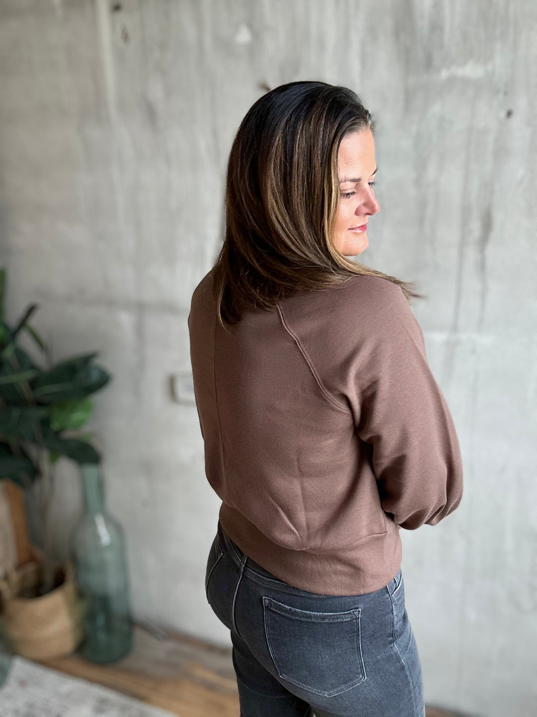 A woman with shoulder-length brown hair is shown from the back wearing a muted brownish-pink cropped sweatshirt and dark gray jeans, standing against a textured concrete wall.
