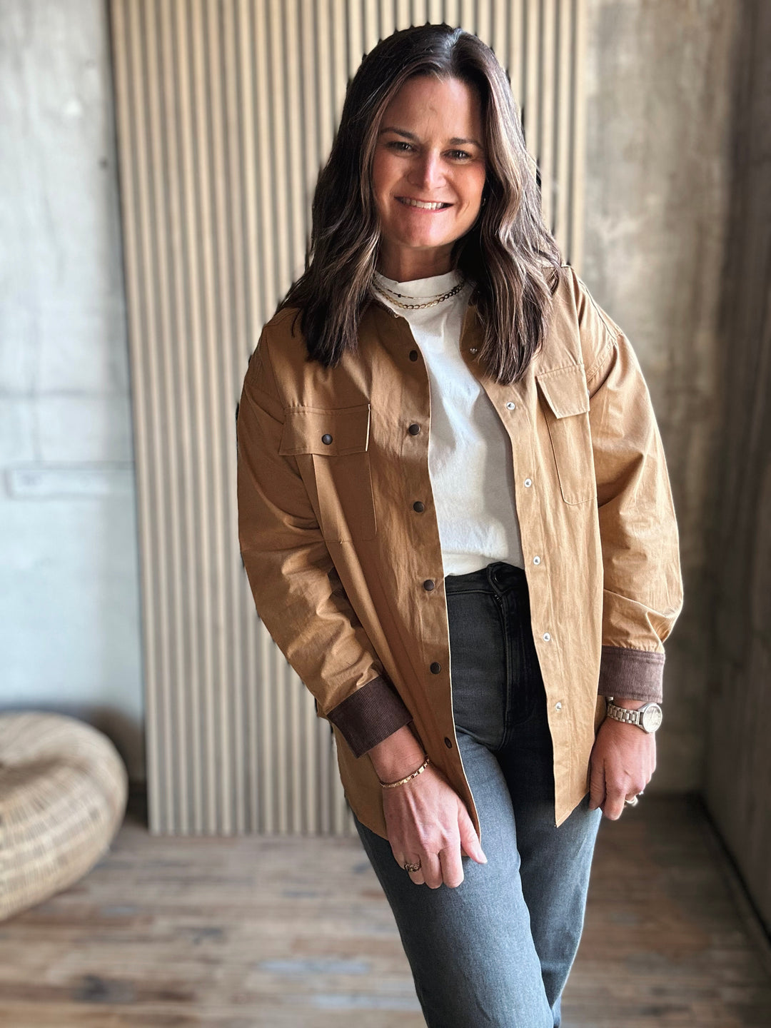 Woman wearing a beige jacket over a white shirt and dark jeans, standing indoors.