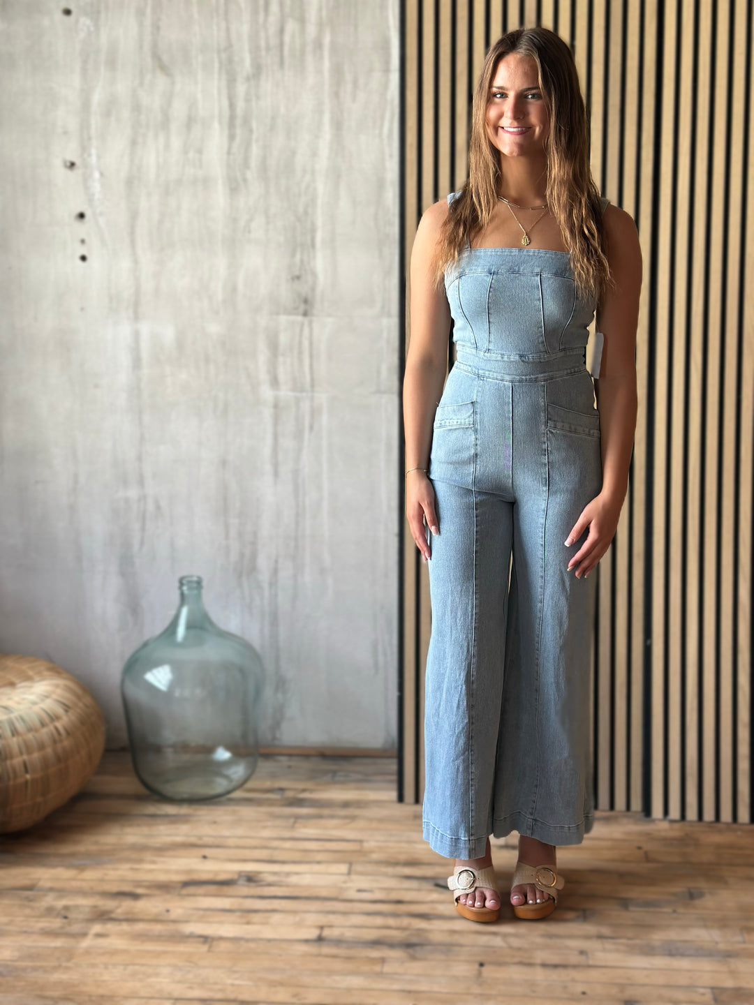 A woman wearing a light blue denim jumpsuit standing in a room with a wooden floor and striped wall.