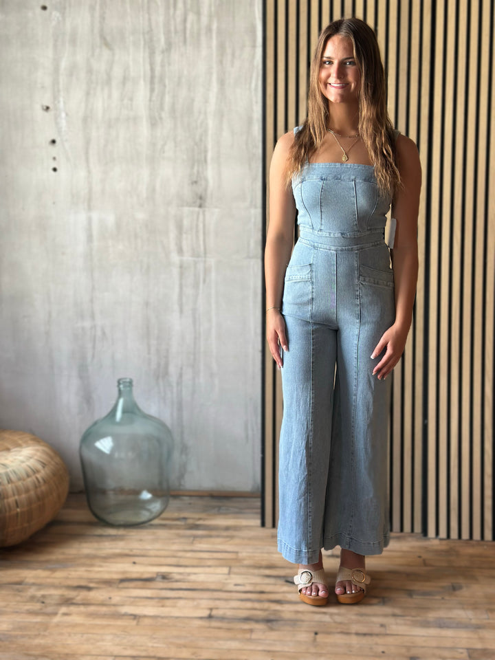 A woman wearing a light blue denim jumpsuit standing in a room with a wooden floor and striped wall.