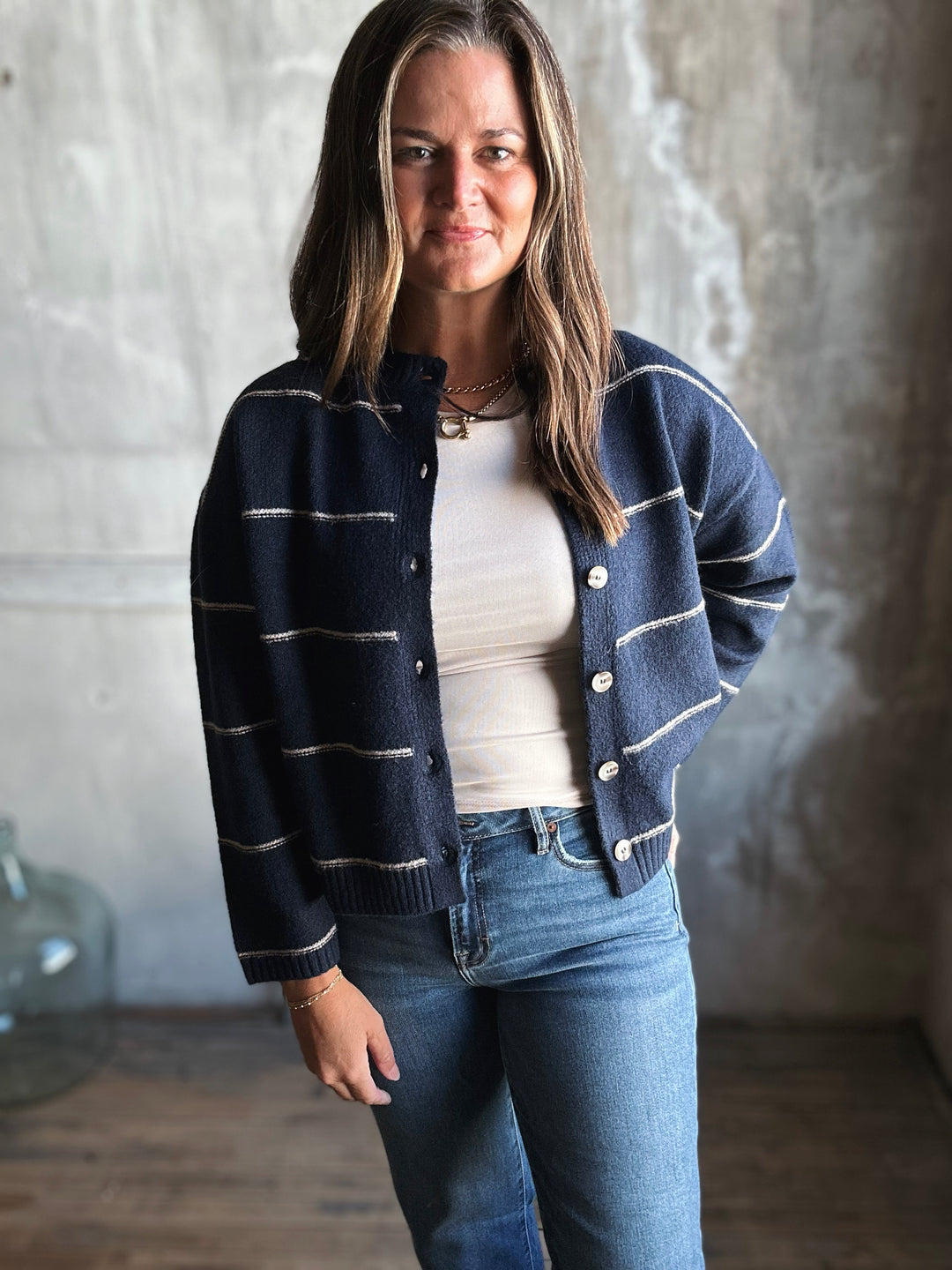 Woman wearing a navy striped cardigan over a white shirt and blue jeans, standing against a textured wall.