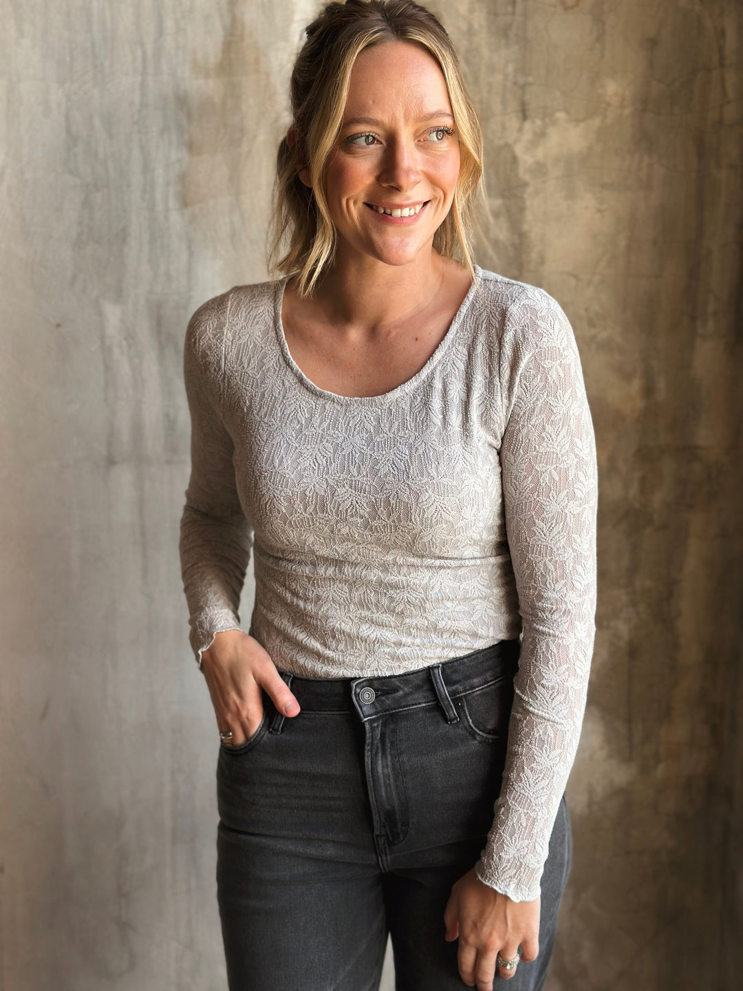 Woman wearing a light gray long-sleeve top and dark jeans against a textured wall.