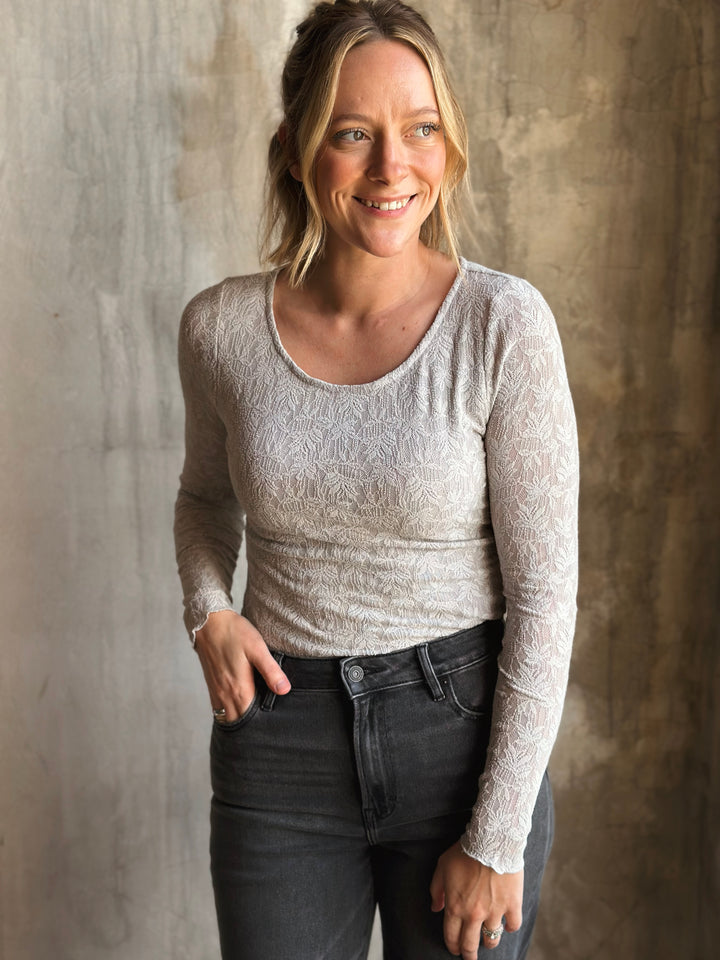 Woman wearing a light gray long-sleeve top and dark jeans against a textured wall.