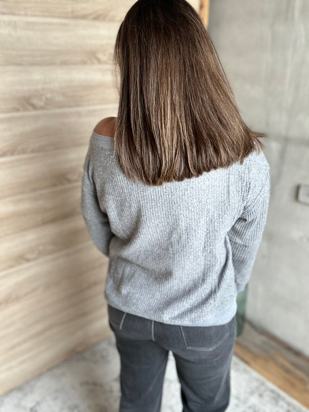 A woman viewed from behind wearing a light gray off-the-shoulder knit sweater and dark gray jeans, standing indoors against a wood-paneled wall.