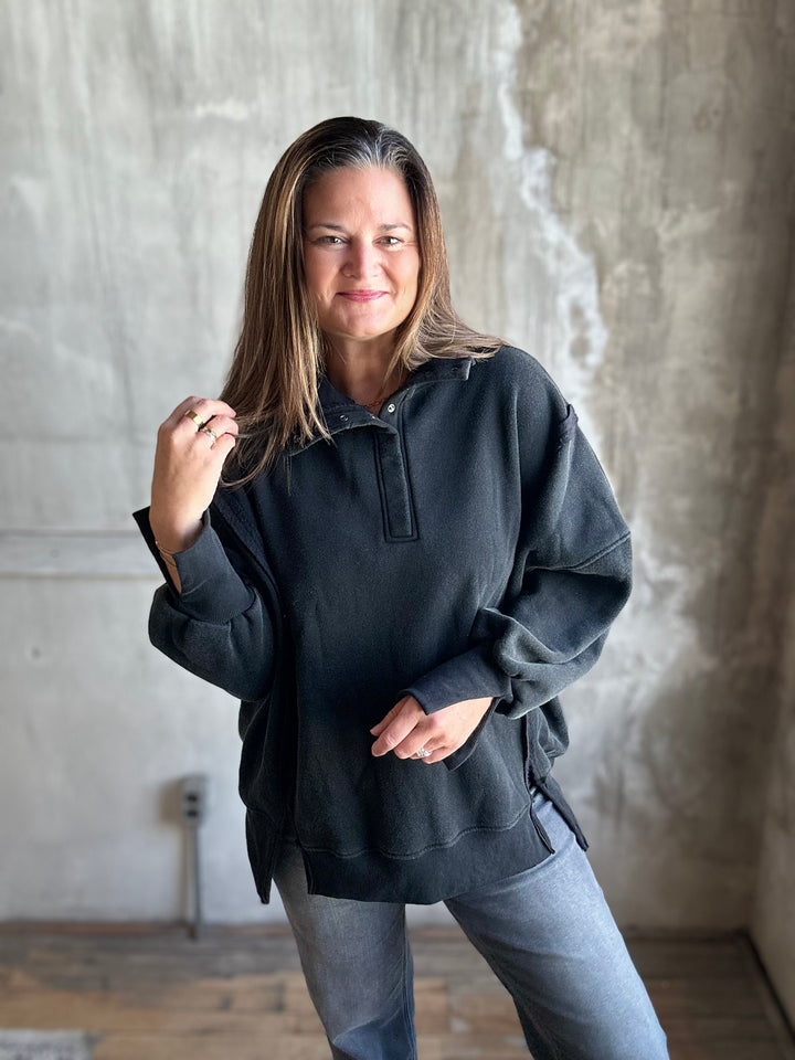 A woman wearing a dark charcoal gray half-placket pullover sweatshirt and light blue jeans, standing in front of a textured concrete wall.