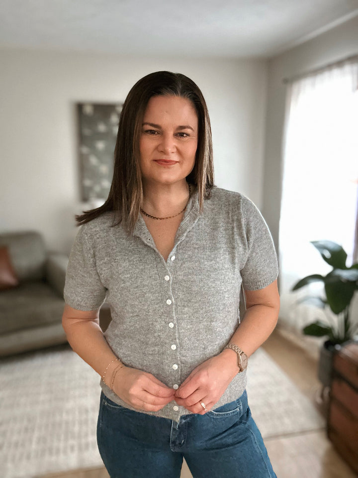 Woman wearing a gray button-up shirt and blue jeans indoors.