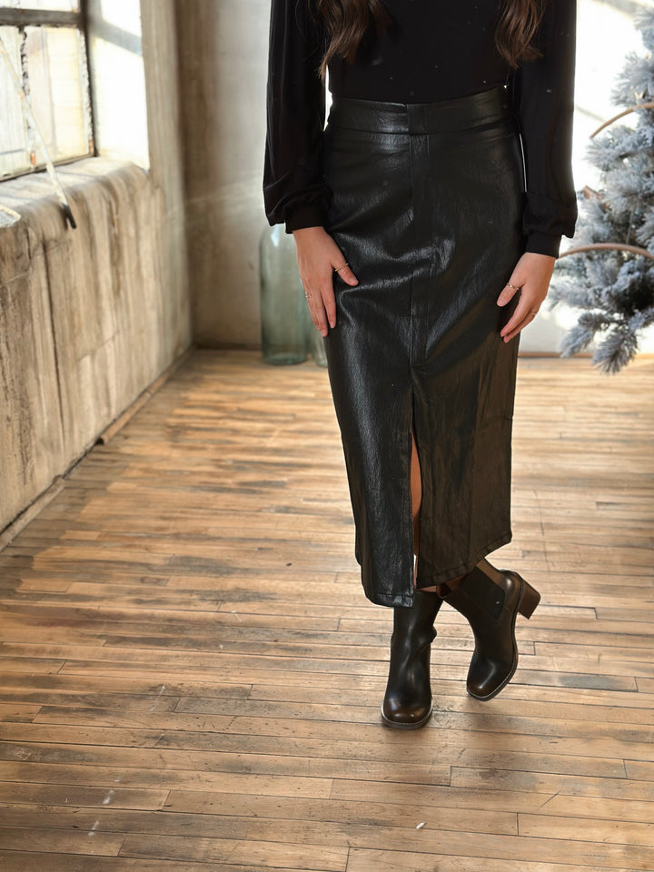 Person wearing a black leather skirt and black boots on a wooden floor.