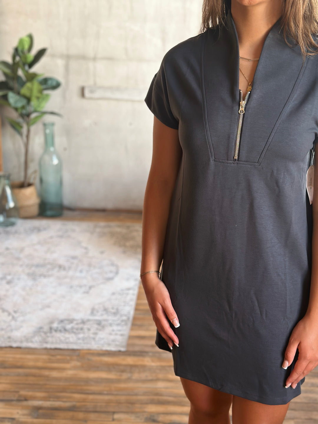 A person wearing a dark gray dress with a zipper in a room with a plant and rug.