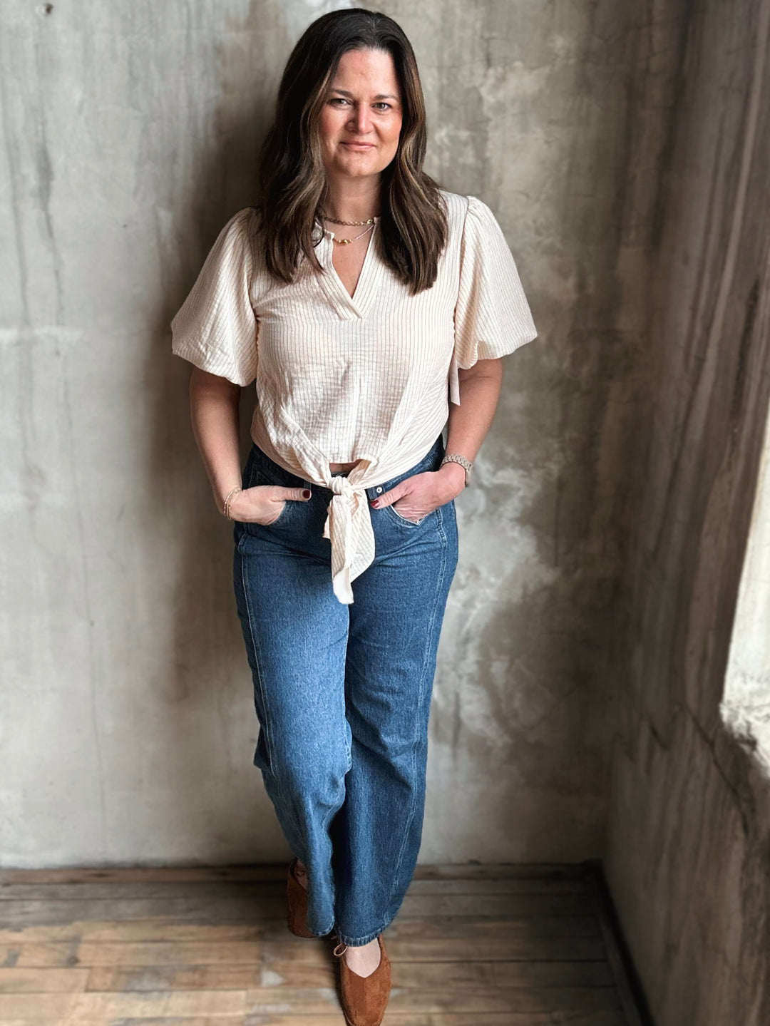 Woman wearing a beige blouse and blue jeans standing against a textured wall.