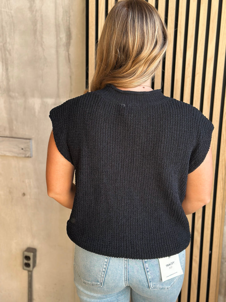 Person wearing a black sleeveless knitted top with light blue jeans, standing against a wooden wall.