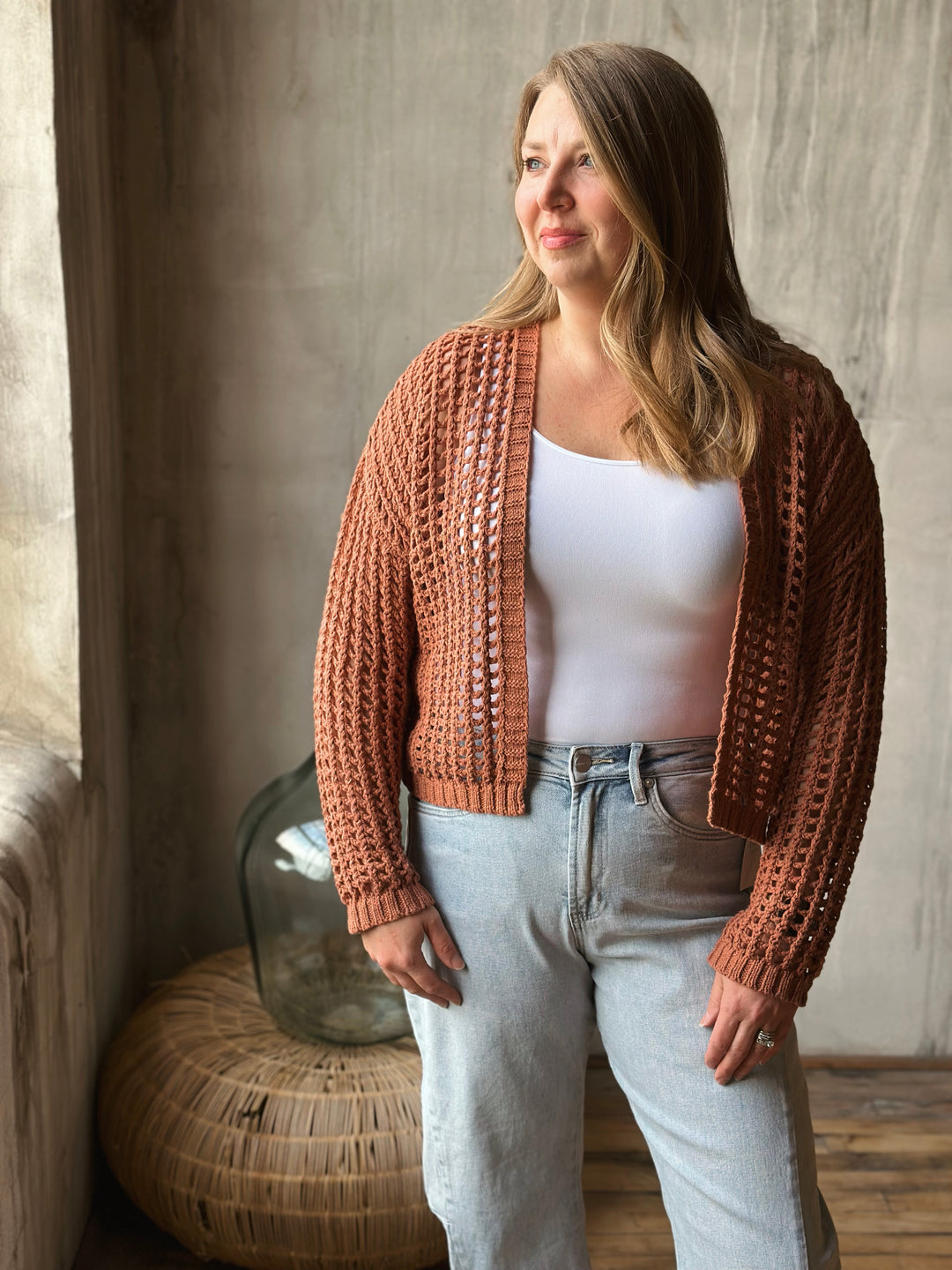 Woman wearing a light brown knitted cardigan over a white top and light blue jeans, standing in a room with a textured wall and wicker basket.