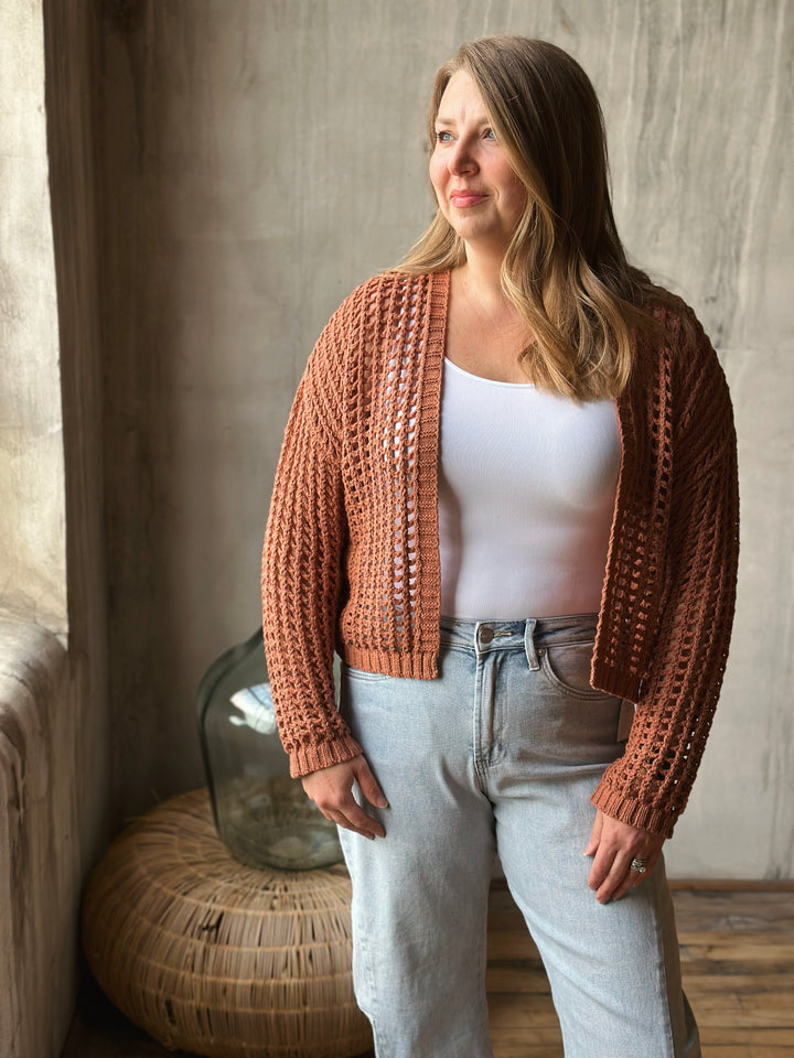 Woman wearing a light brown knitted cardigan over a white top and light blue jeans, standing in a room with a textured wall and wicker basket.