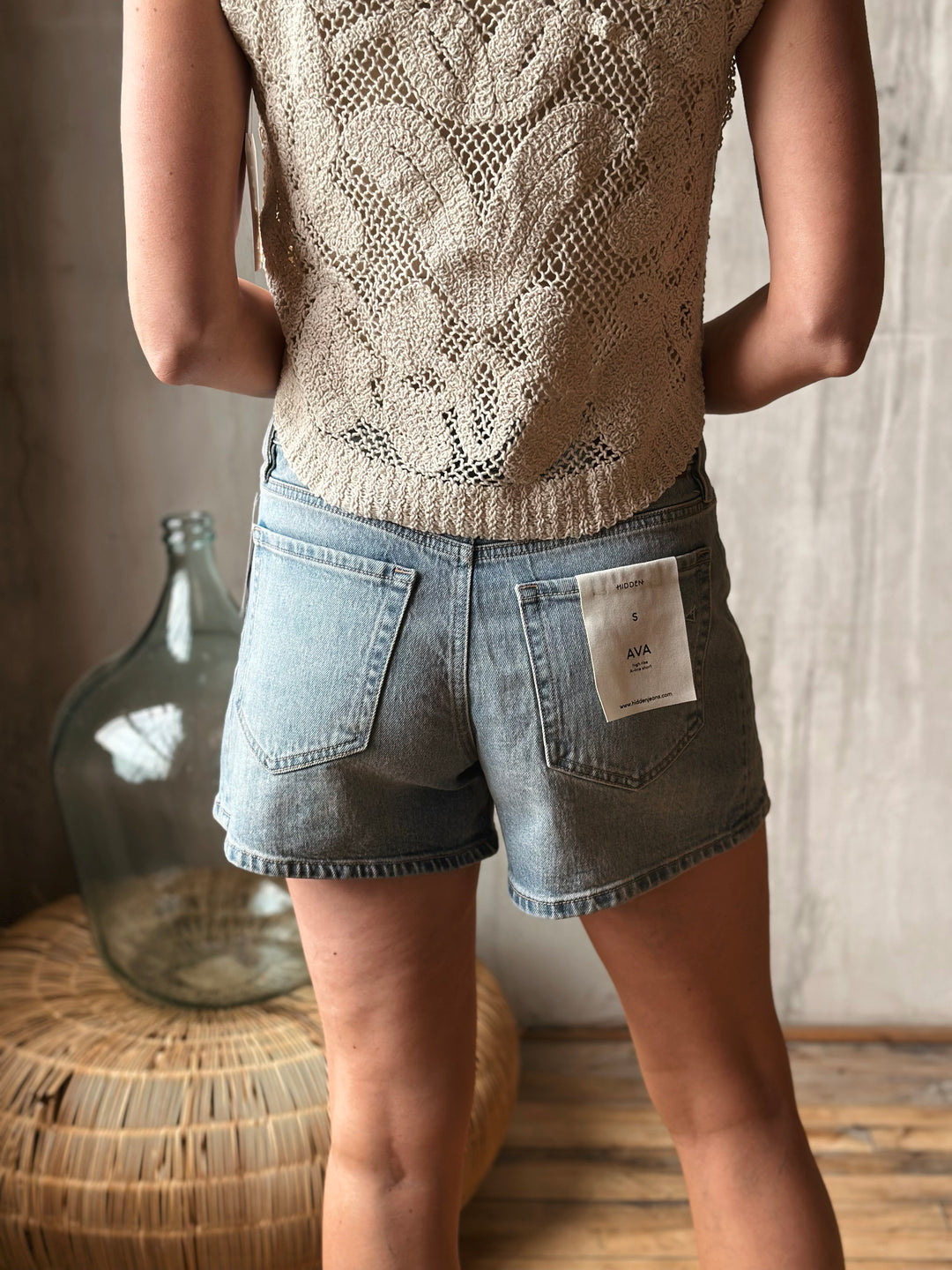 A person wearing a beige knitted top and light washed denim shorts standing indoors.
