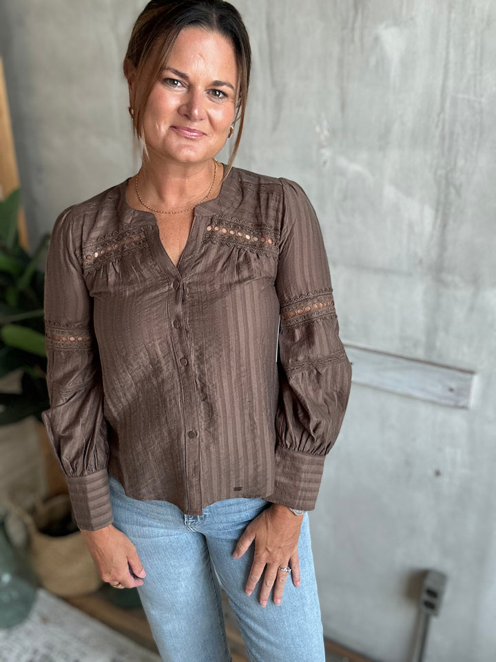 A woman wearing a brown long-sleeve button-up blouse with vertical stripes and lace-up detailing, paired with light blue jeans, standing against a textured gray wall.