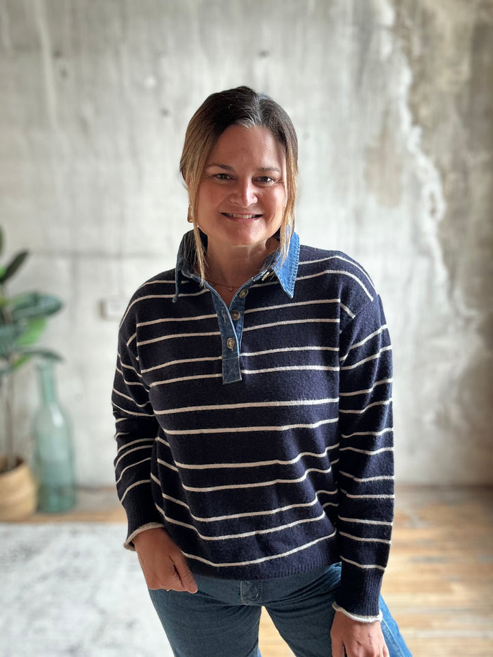 A woman wearing a navy blue and white striped long-sleeve knit polo sweater with a blue denim collar, standing in a room with a textured concrete wall background.