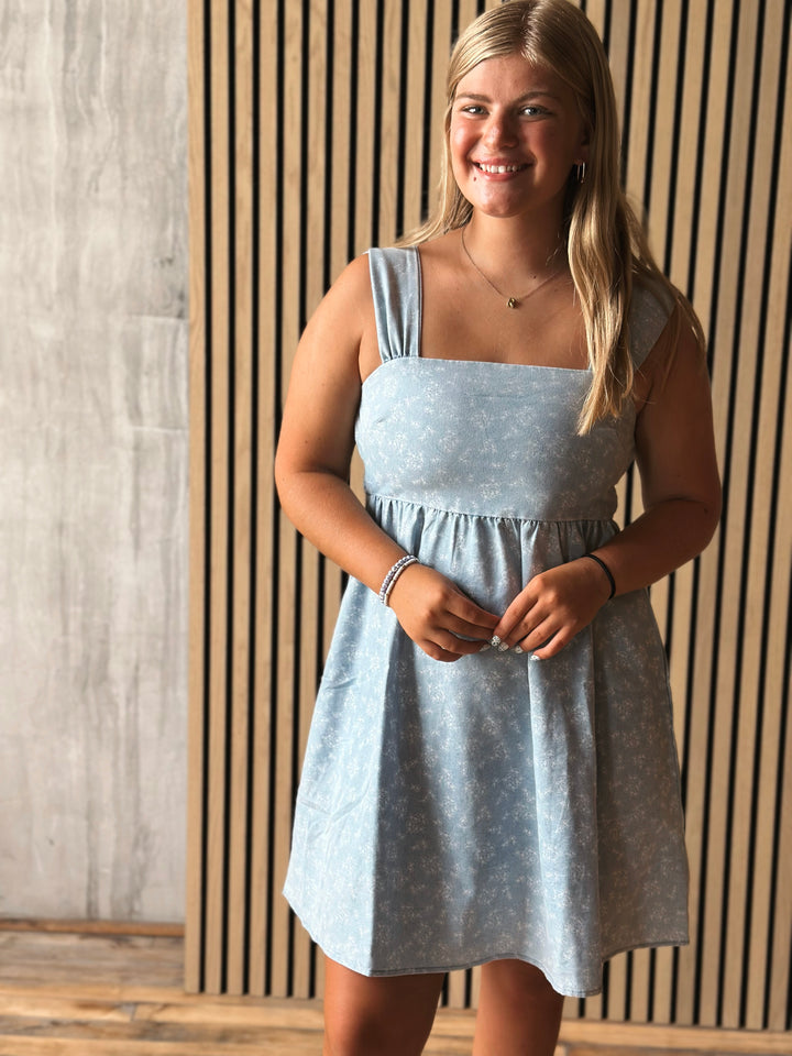Woman wearing a light blue dress standing against a wooden paneled wall.