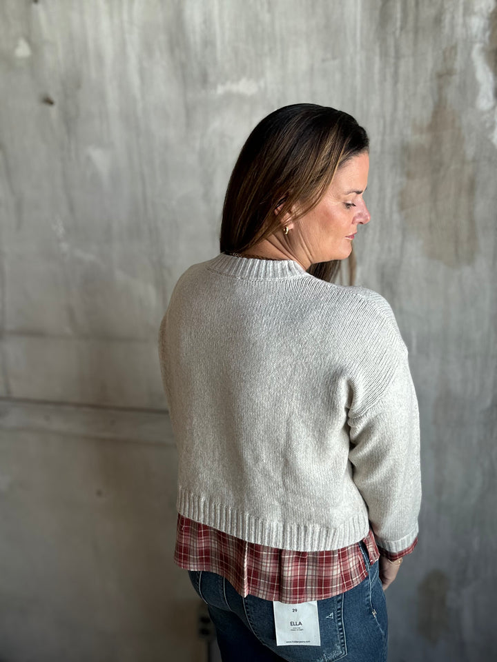 A woman is shown from the back wearing a light gray ribbed knit sweater, a red and white plaid shirt, and blue jeans, standing against a textured concrete wall.