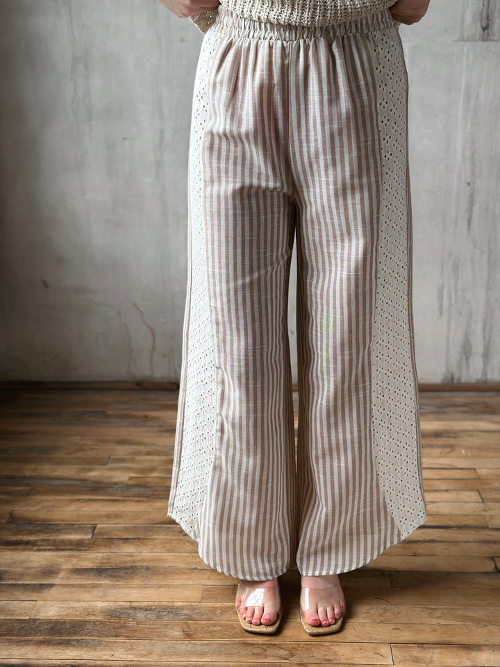 Person wearing beige striped pants with lace detail on the side standing against a wooden floor and gray wall.