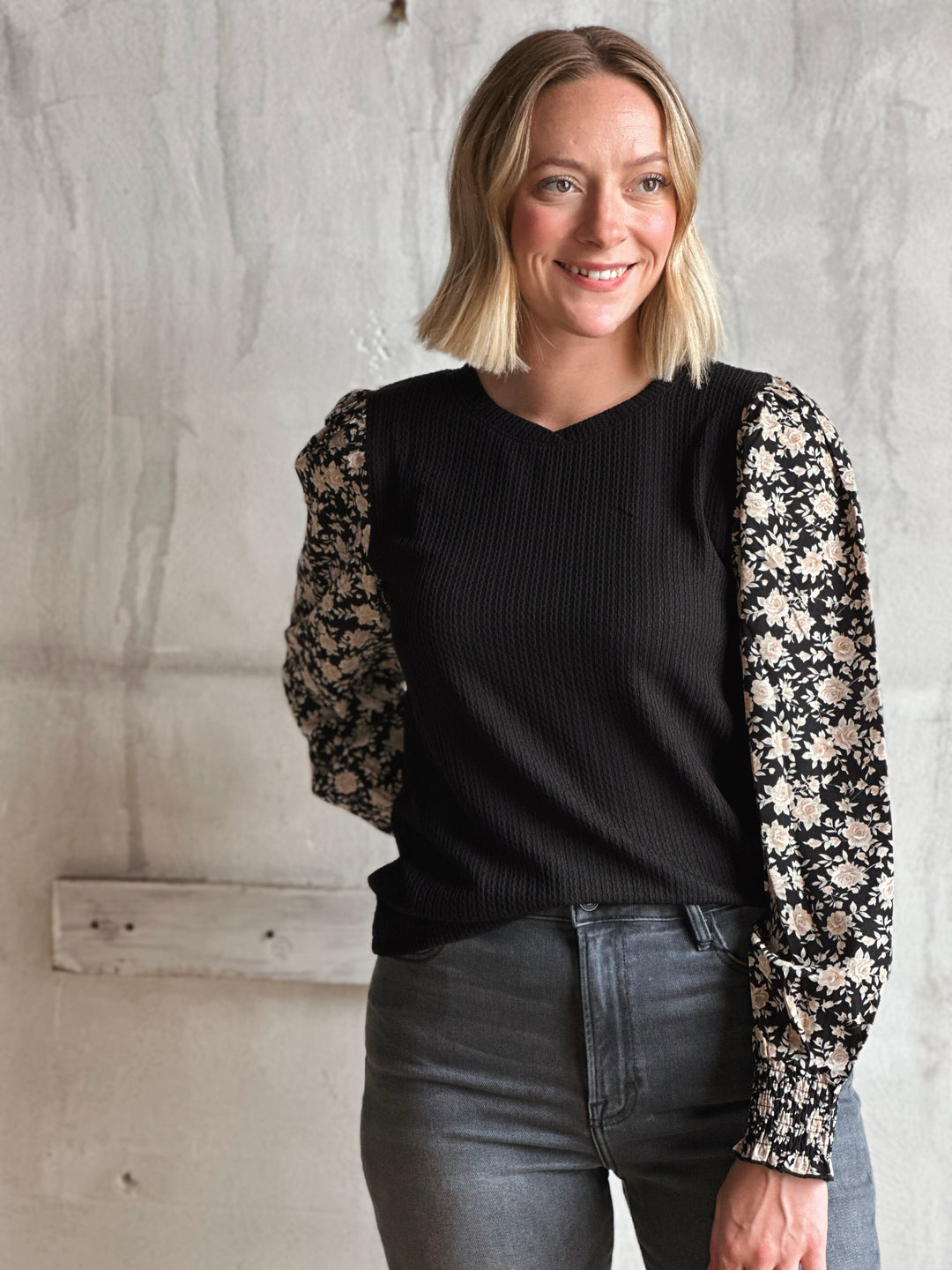 Woman wearing a black sweater with floral sleeves against a textured wall.