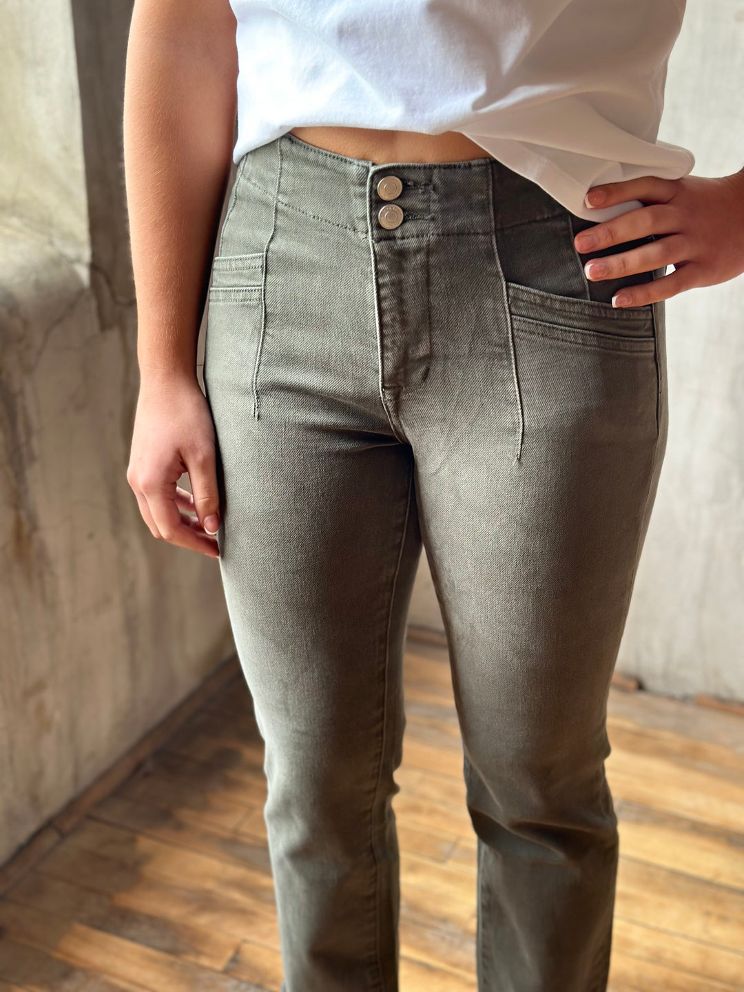 A person wearing slim-fit gray jeans with a high waist and double button closure, paired with a white cropped top, standing in an indoor setting with a concrete wall and wooden floor.