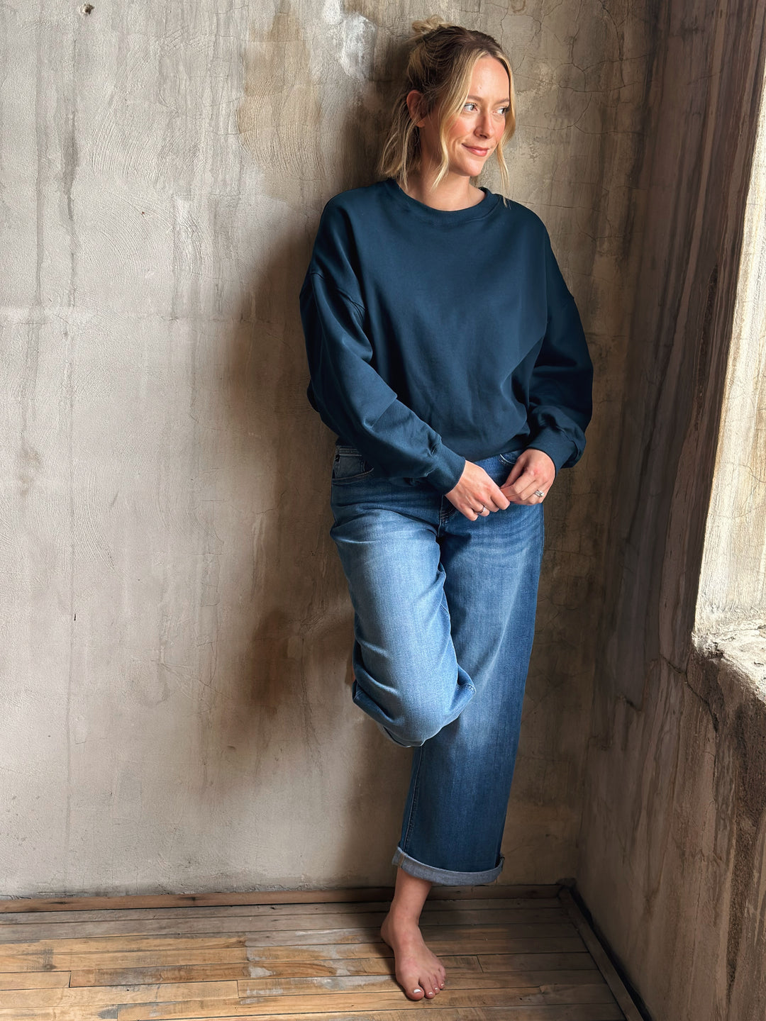 Person wearing a blue sweatshirt and jeans leaning against a textured wall.