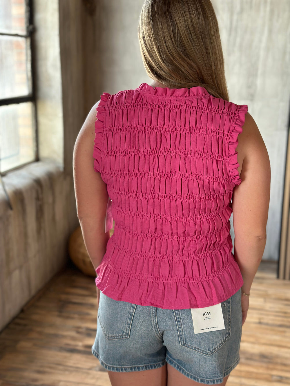 A person wearing a pink ruffle sleeveless top and denim shorts in a room with a wooden floor and large window.