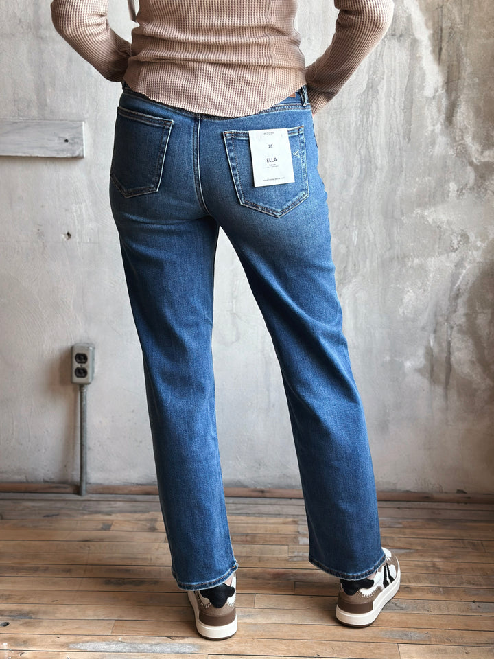 Person wearing blue jeans with a beige sweater and white sneakers in a room with wooden floor and gray wall.