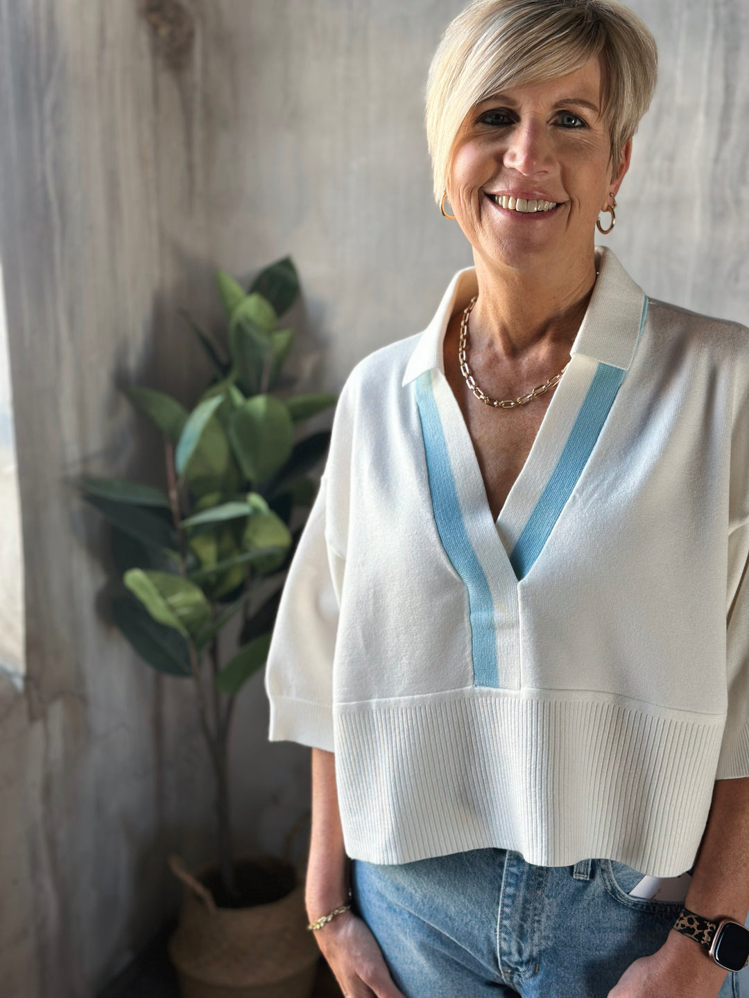 Woman wearing a white blouse with blue accents standing indoors next to a plant.