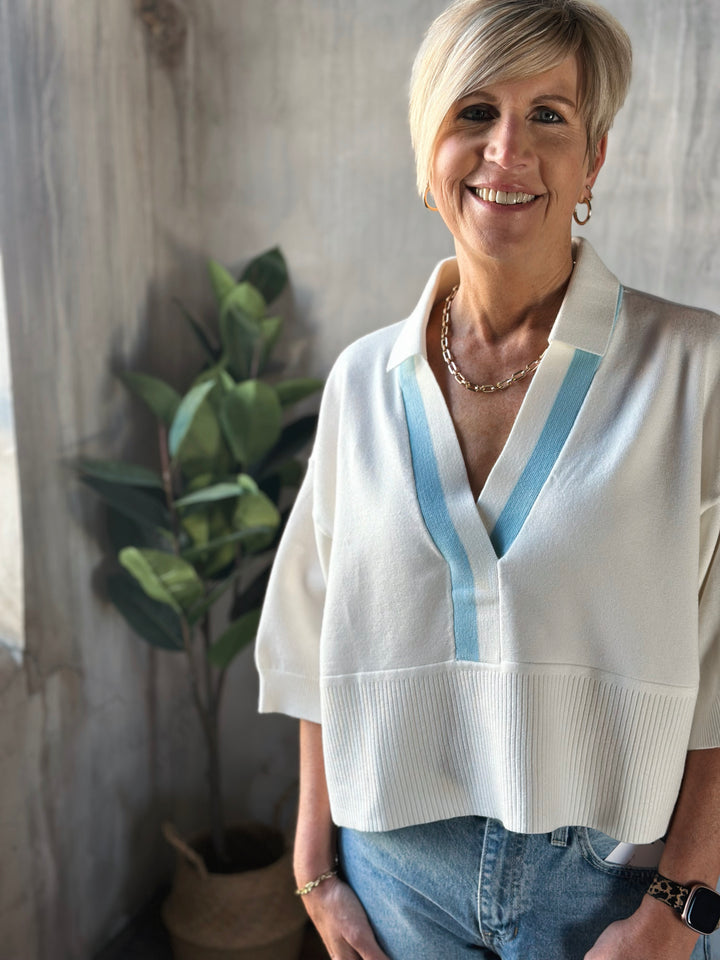Woman wearing a white blouse with blue accents standing indoors next to a plant.