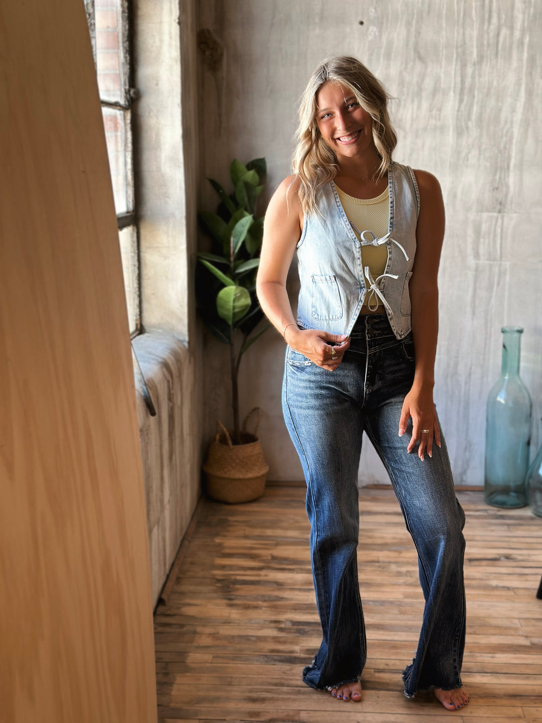 Woman wearing a sleeveless denim vest and flared jeans standing in a room with wooden floor and light-colored walls.