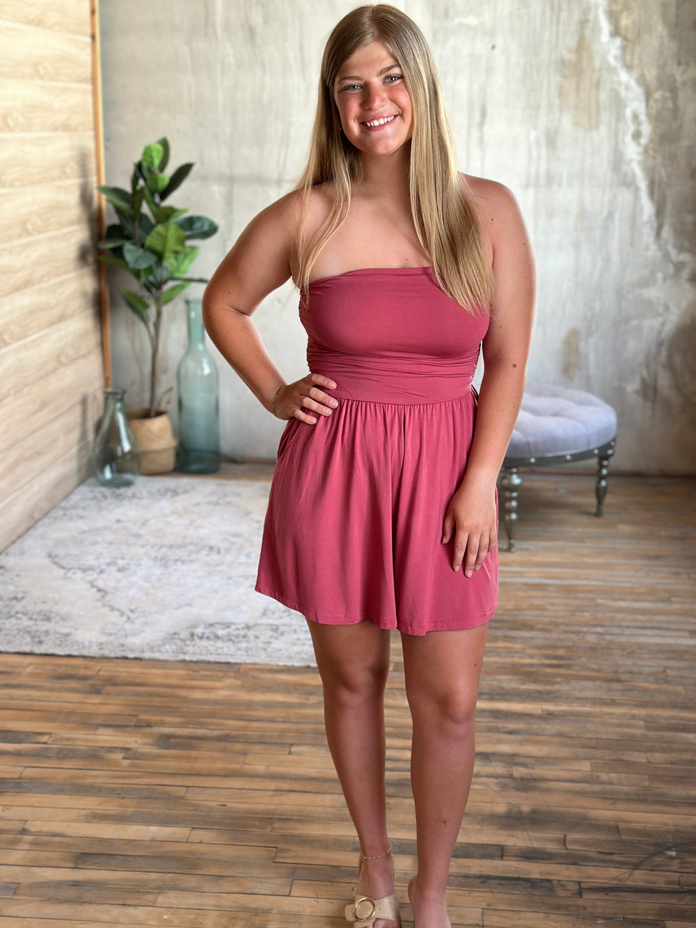 Woman wearing a pink strapless romper standing in a room with wooden floor and plant decor.