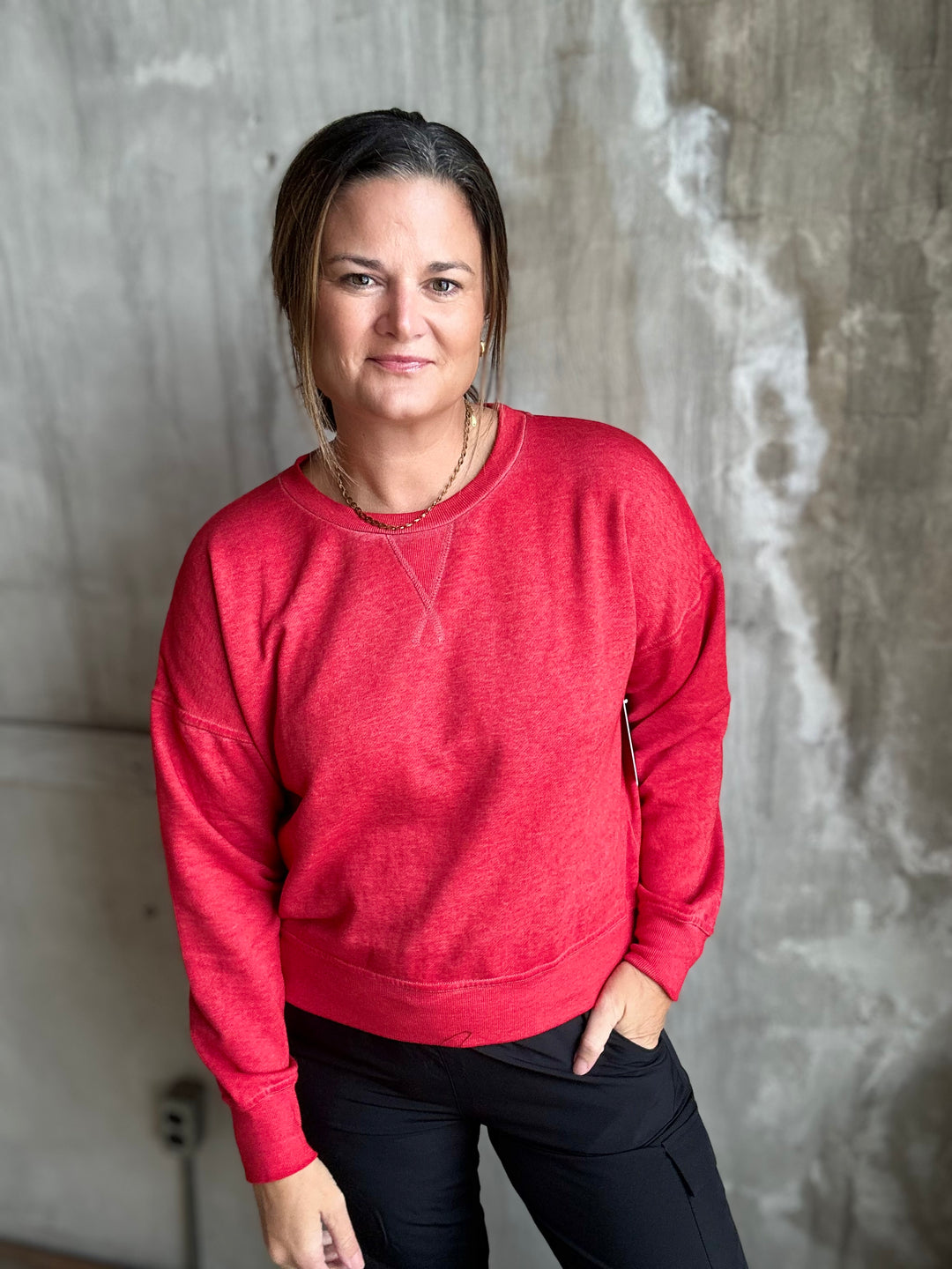 A woman wearing a heather red crewneck sweatshirt and black pants, standing against a textured gray concrete wall.