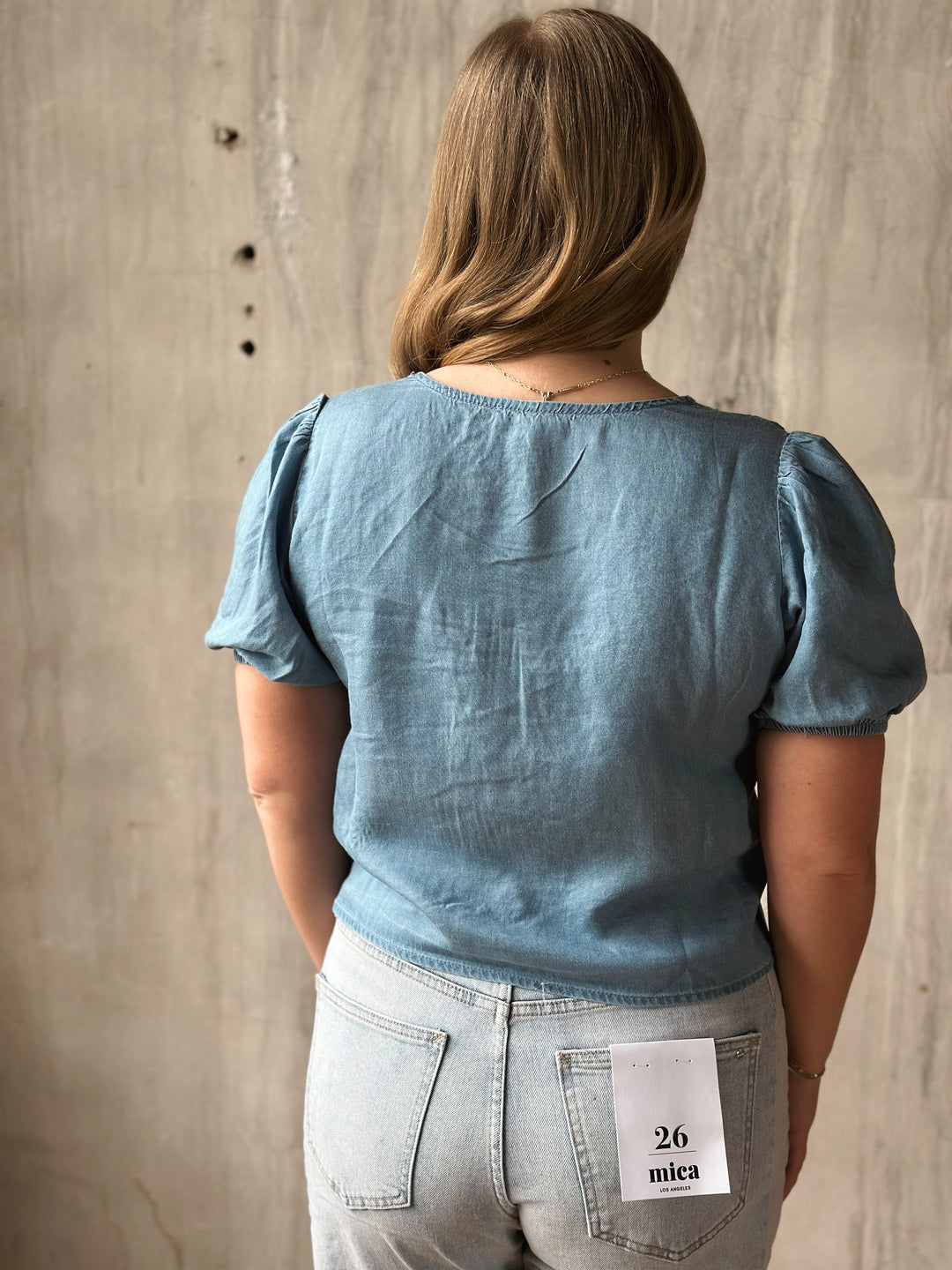 Person wearing a blue top and light blue jeans with a visible brand label against a concrete wall.