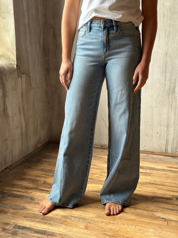 A person wearing light-wash wide-leg denim jeans and a white top, standing barefoot on a wooden floor against a textured concrete wall.