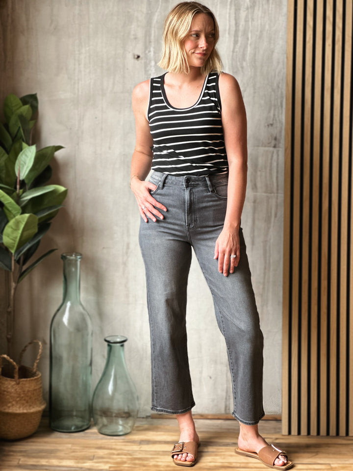 Woman wearing a black and white striped tank top and gray jeans standing against a wooden paneled wall.