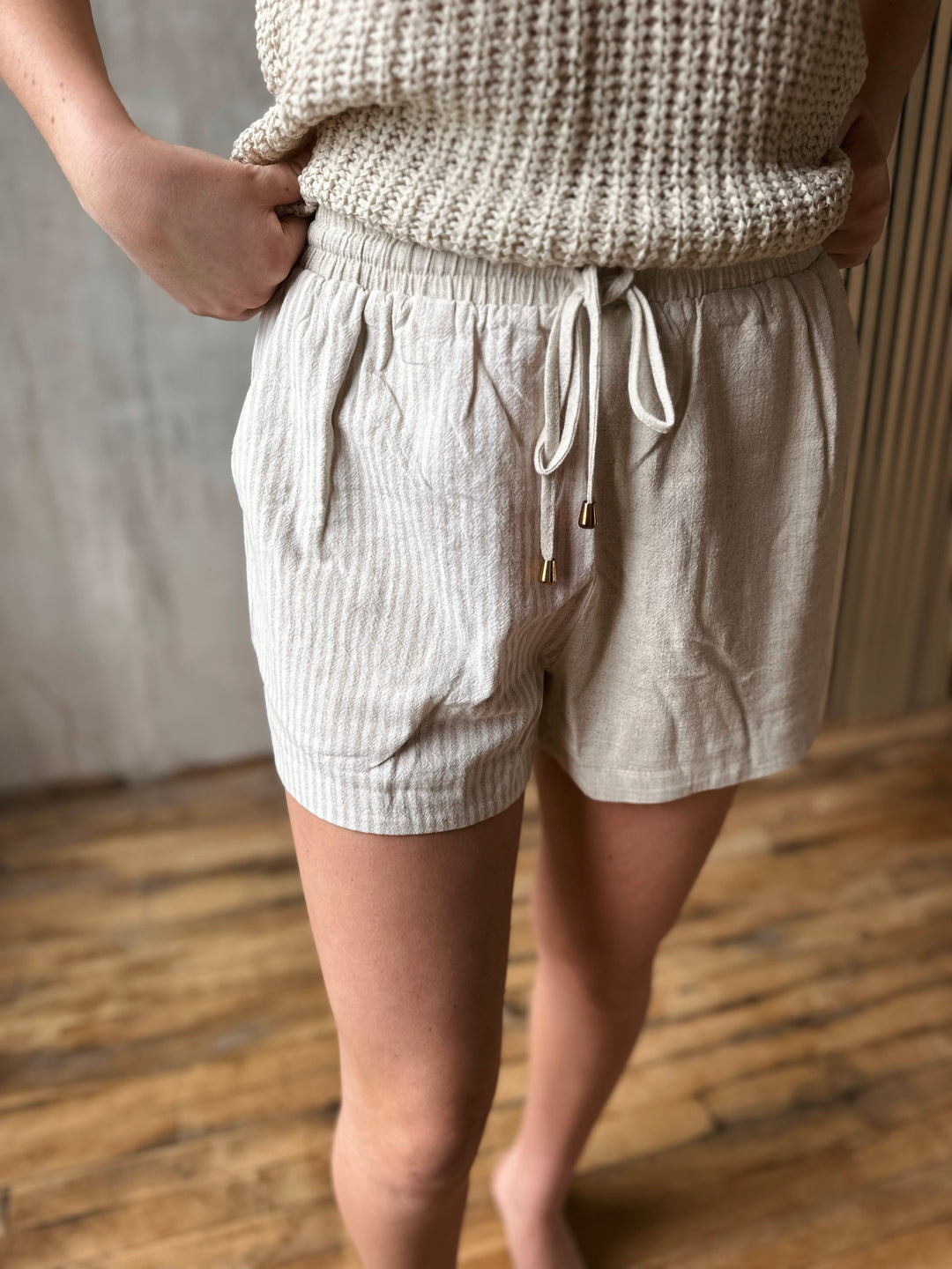 Person wearing beige shorts with one half having stripes and a beige knitted top on a wooden floor.