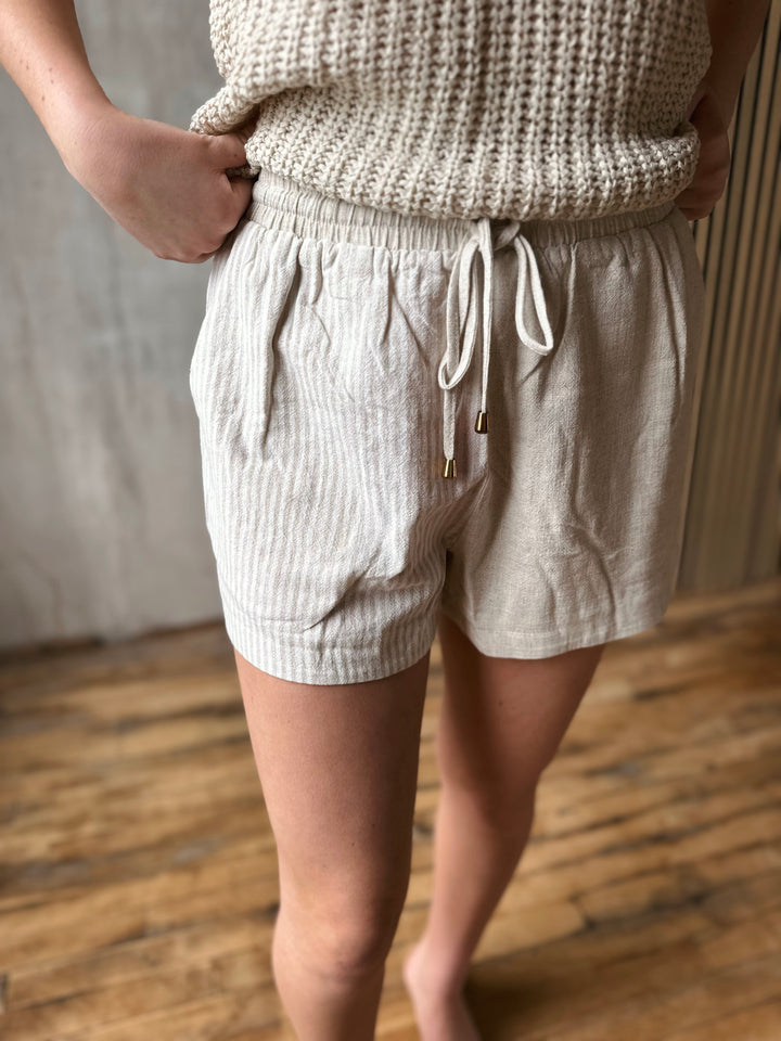 Person wearing beige shorts with one half having stripes and a beige knitted top on a wooden floor.