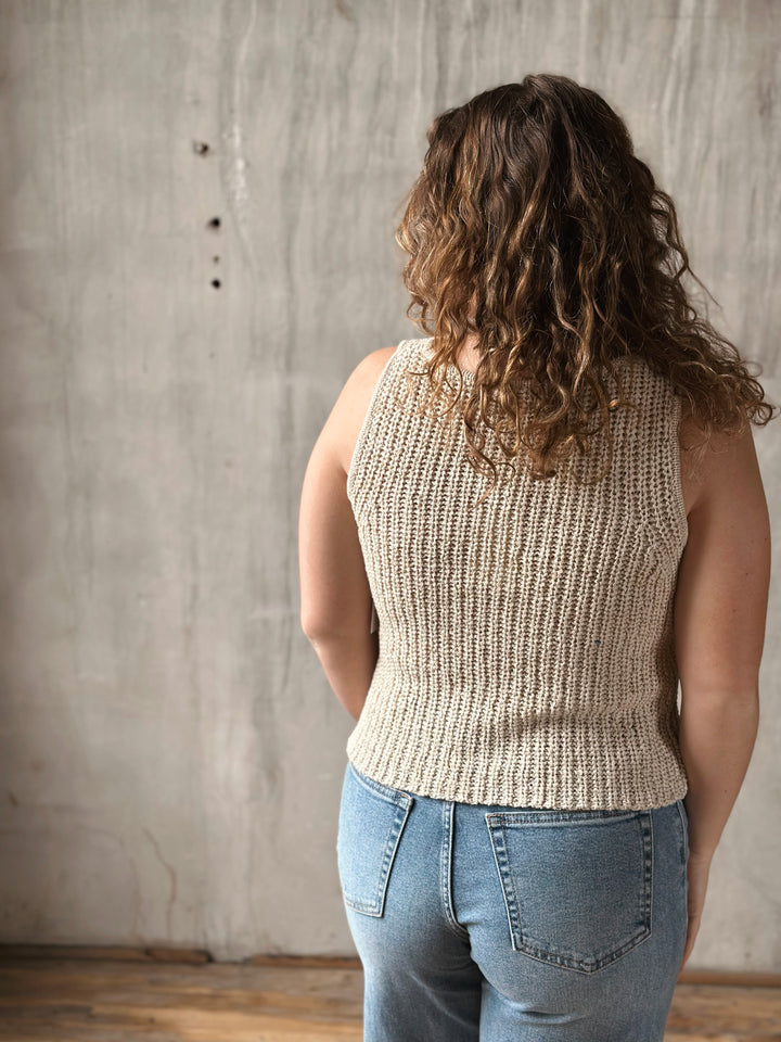 Person wearing a beige knitted sleeveless top and blue jeans against a textured wall.