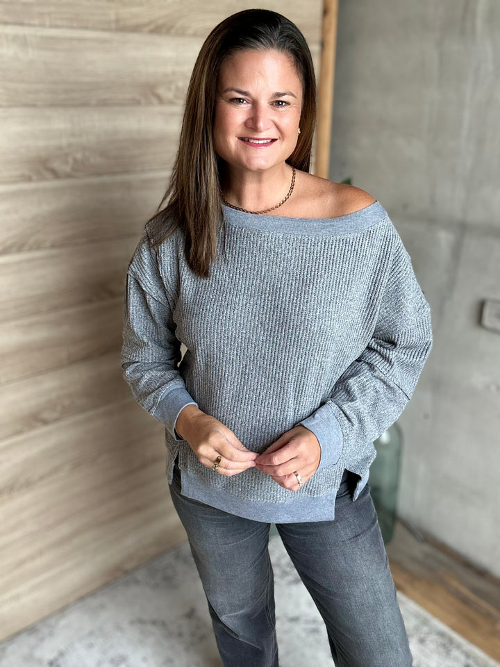 A woman wearing a light gray off-the-shoulder ribbed knit sweater and dark gray jeans, standing in an indoor setting with a wood-paneled wall and concrete wall in the background.