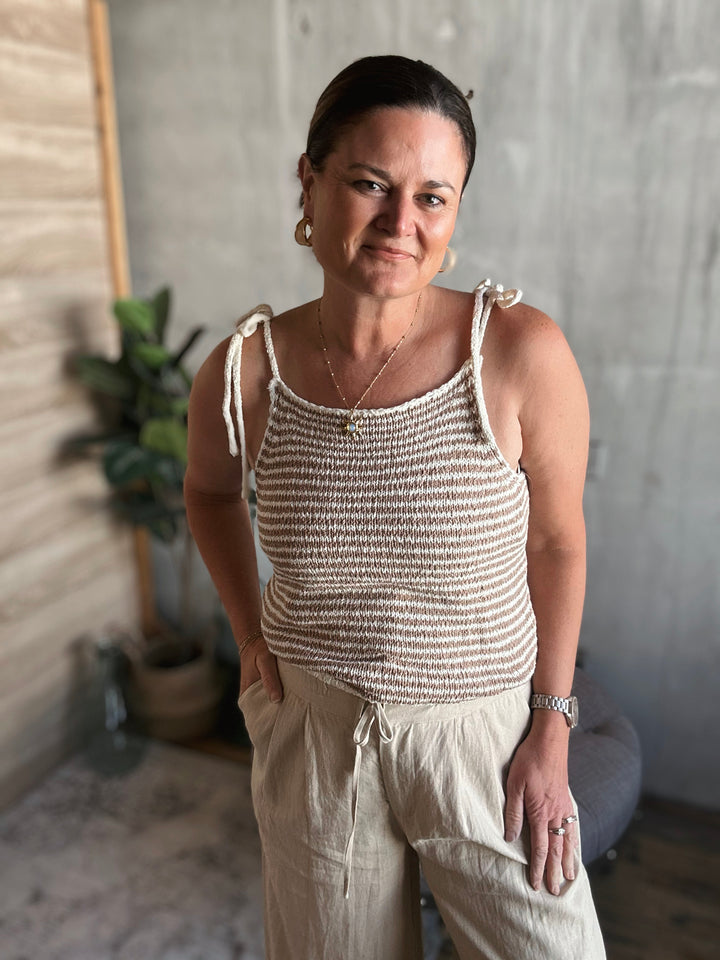 A woman wearing a light brown and beige striped crocheted tank top with thin straps that are tied into small bows.