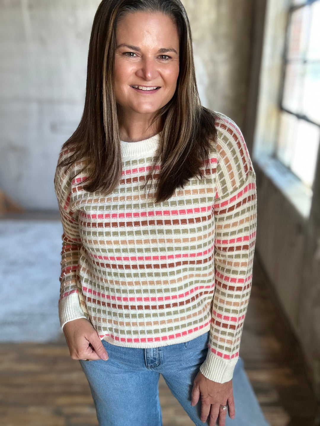 Woman wearing a cream crewneck sweater with pink, green and red stripes and light-wash jeans