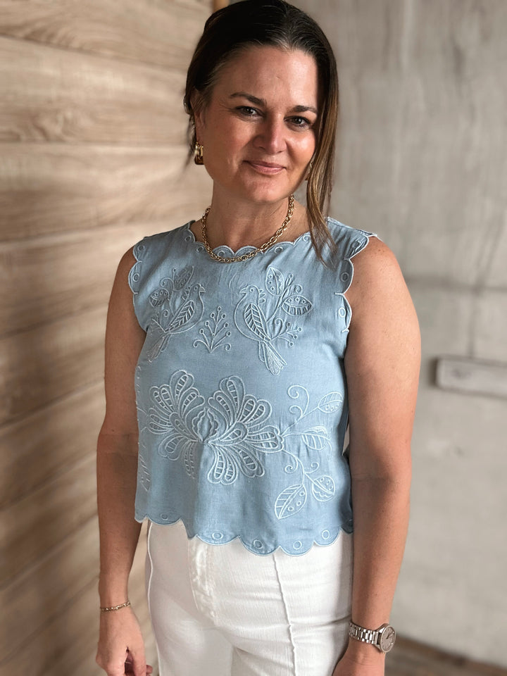 Woman wearing a light blue embroidered top with white pants against a neutral wall.