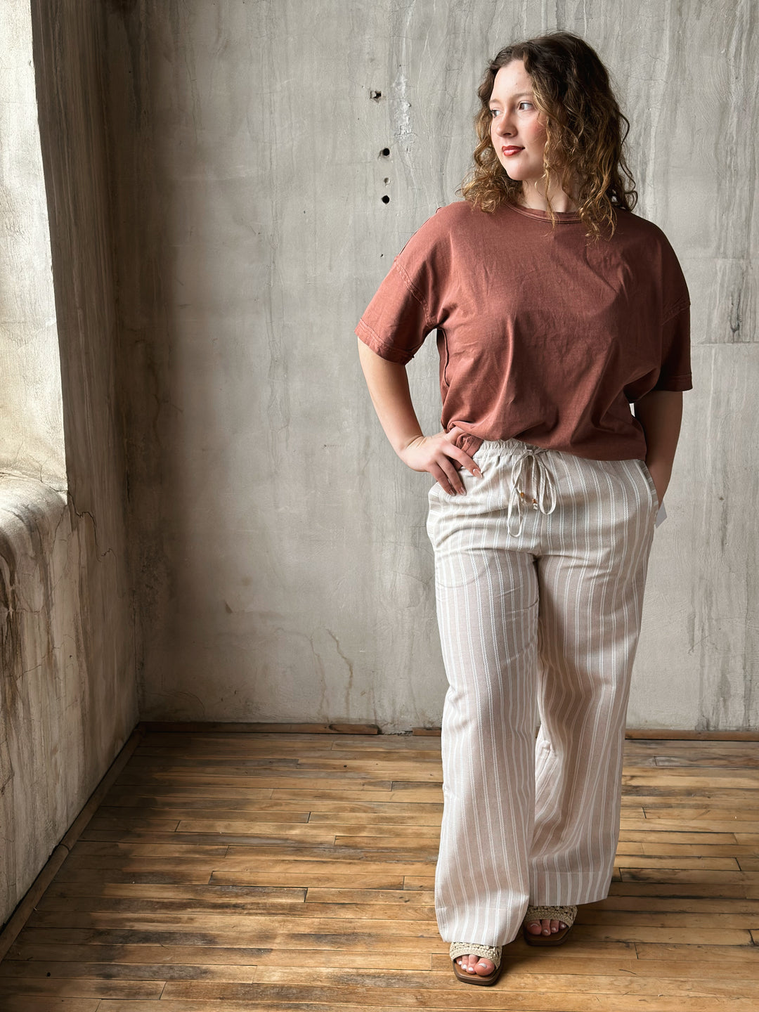 Woman wearing a brown top and white striped pants standing in a room with wooden floor and concrete wall.