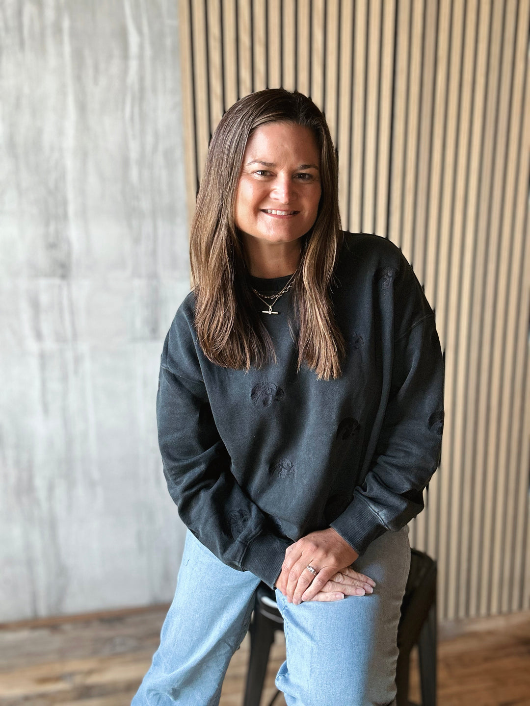Woman wearing a dark gray sweatshirt and light blue jeans sitting on a stool against a striped wall.