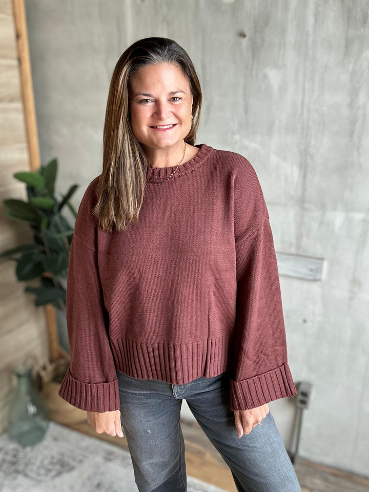 A woman wearing a dusty rose crewneck knit sweater with dropped shoulders and ribbed cuffs, paired with gray jeans, standing in front of a textured concrete wall.