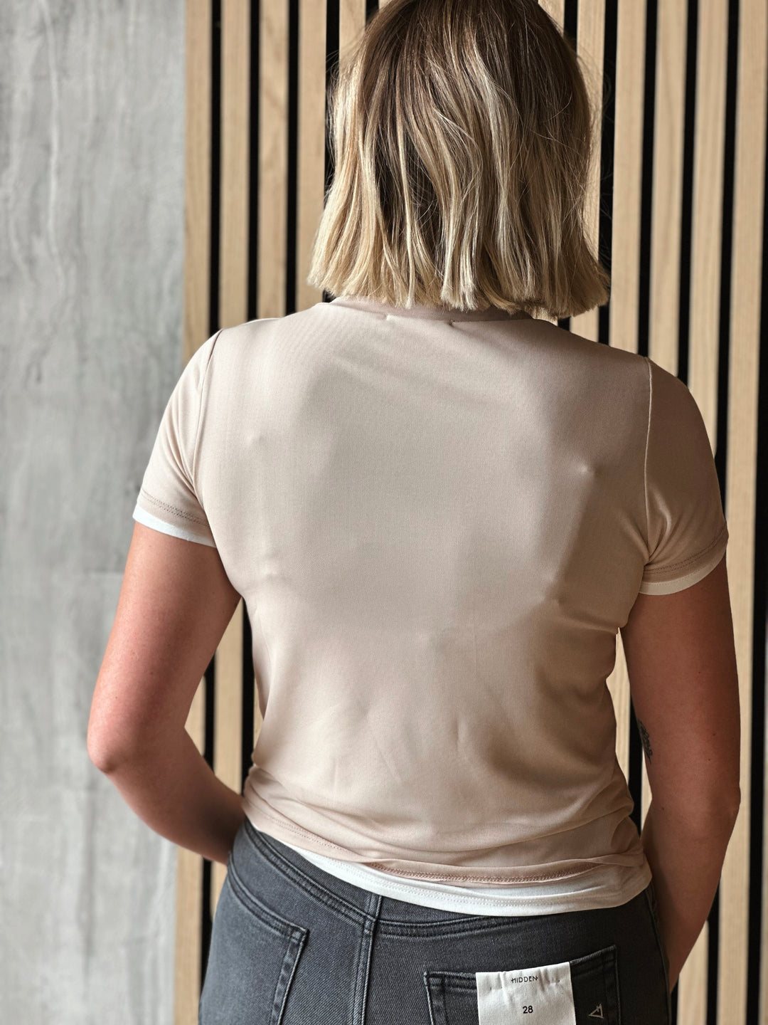 Person wearing a beige t-shirt and gray pants against a wooden panel background